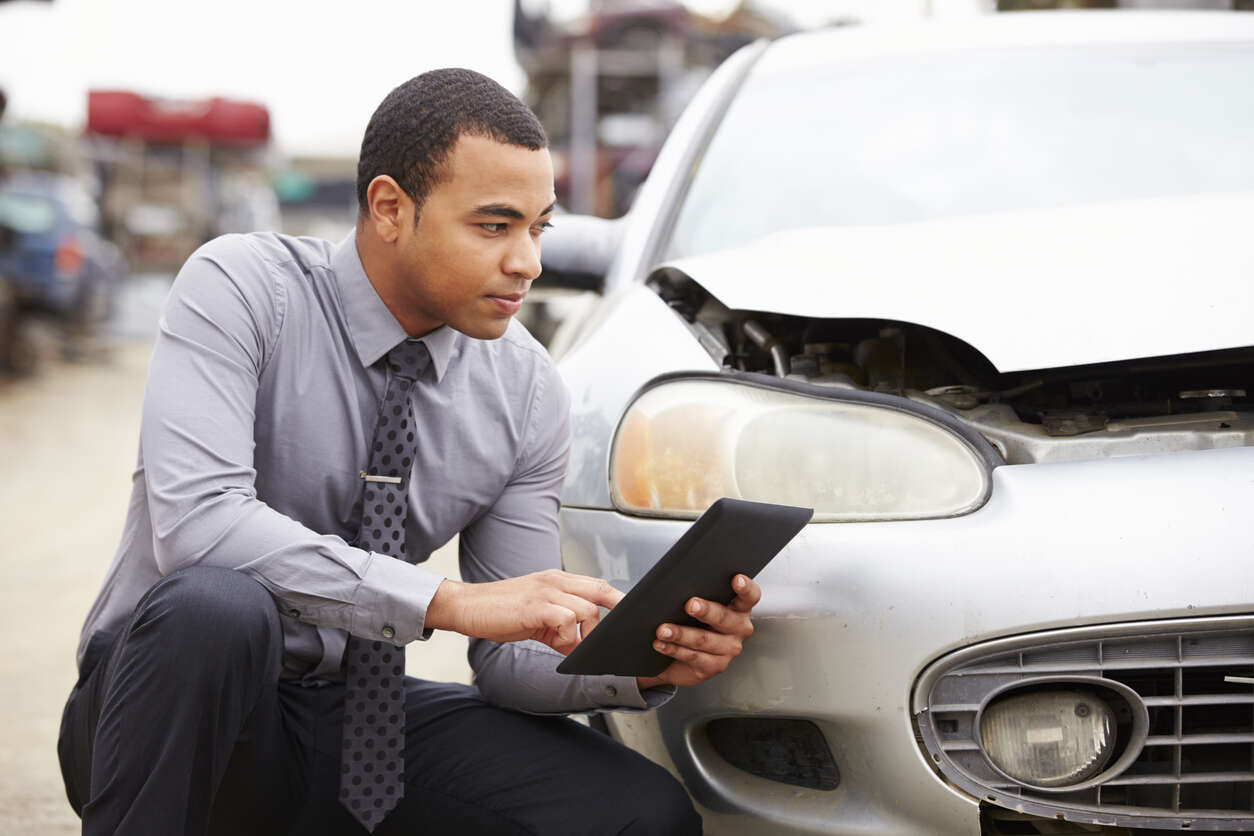 A man in a suit examines a damaged car with a tablet, symbolizing the investigation of auto claims when injured in a company car in California.